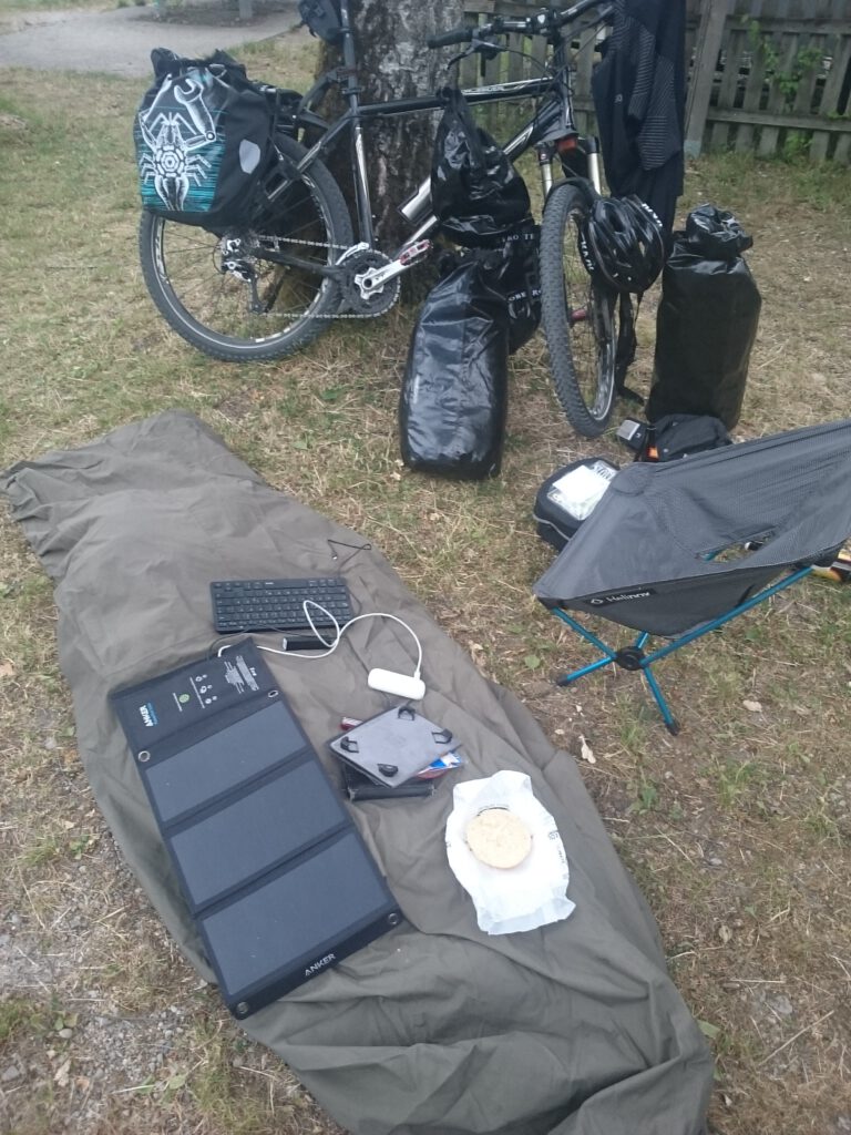 Mein erstes Nachtlager: Im Vordergrund mein Biwaksack, daneben ein Campinghocker und im Hintergrund mein Fahrrad an einen Baum gelehnt.
Auf dem Biwaksack liegt eine Solarzelle zum Laden diverser elektronischer Geräte.